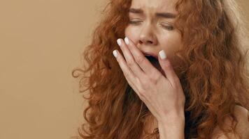 A young woman with curly red hair yawns broadly, displaying a moment of tiredness. She stands against a simple, light-colored backdrop, capturing the essence of fatigue. video