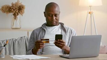 A man in his 30s sits comfortably in a modern home setting, using his smartphone while holding a credit card. He is focused on managing his finances in a relaxed atmosphere. video