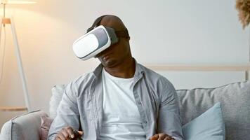 A young African American man engages with a virtual reality headset while sitting on a comfortable sofa in a stylish living room. He is focused and enjoying the gaming moment. video