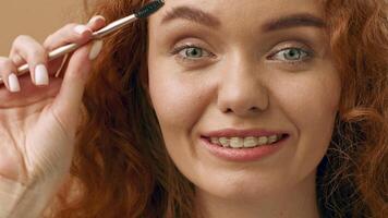 A young woman with curly red hair smiles while using a brow brush. The bright interior enhances her cheerful expression, showcasing her beauty routine in a relaxed environment. video