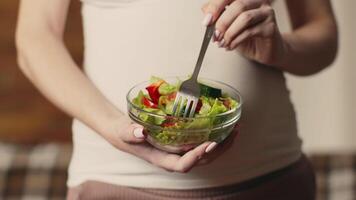 A pregnant woman holds a bowl of fresh salad, fork in hand, savoring healthy greens and vegetables while relaxing in her cozy home during the afternoon. video