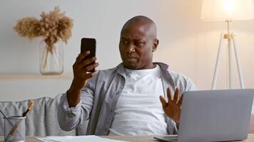 A man in his 30s with a focused expression looks at his smartphone while seated at a home office desk. A laptop and notepad are nearby, and warm light enhances the cozy atmosphere. video