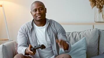 African American man is sitting on a comfortable couch and playing games, enjoying his time at home in a relaxed atmosphere. Natural light fills the space. video