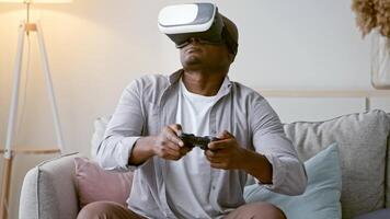 A young African American man in his 30s immerses himself in a virtual reality gaming experience. He sits comfortably on a sofa, wearing a VR headset and holding a controller, in a stylish living room. video