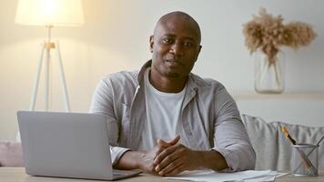 A man in his 30s sits comfortably at his desk in a stylish living space, engaging with the camera while surrounded by documents and a laptop. Natural light fills the room, creating a warm atmosphere. video