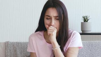 A young woman shows signs of illness, coughing while sitting at home. She appears unwell and is likely suffering from flu, cold, or other respiratory issues in isolation. video