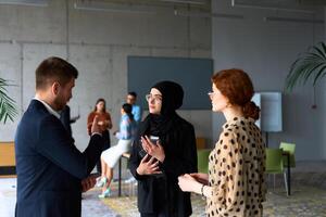 A diverse group of business professionals engages in a conversation during a work break in the office, with their colleagues visible in the background, fostering collaboration and connection photo