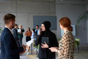 A diverse group of business professionals engages in a conversation during a work break in the office, with their colleagues visible in the background, fostering collaboration and connection photo