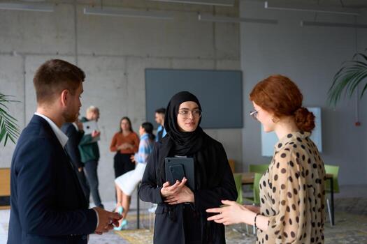 A diverse group of business professionals engages in a conversation during a work break in the office, with their colleagues visible in the background, fostering collaboration and connection photo