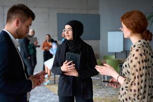 A diverse group of business professionals engages in a conversation during a work break in the office, with their colleagues visible in the background, fostering collaboration and connection photo