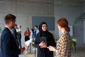 A diverse group of business professionals engages in a conversation during a work break in the office, with their colleagues visible in the background, fostering collaboration and connection photo