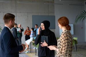 A diverse group of business professionals engages in a conversation during a work break in the office, with their colleagues visible in the background, fostering collaboration and connection photo