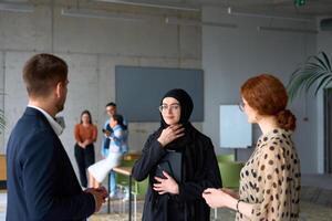 A diverse group of business professionals engages in a conversation during a work break in the office, with their colleagues visible in the background, fostering collaboration and connection photo