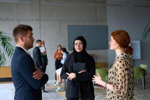 A diverse group of business professionals engages in a conversation during a work break in the office, with their colleagues visible in the background, fostering collaboration and connection photo