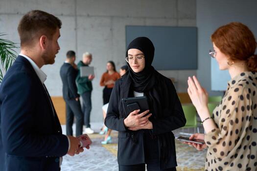 A diverse group of business professionals engages in a conversation during a work break in the office, with their colleagues visible in the background, fostering collaboration and connection photo