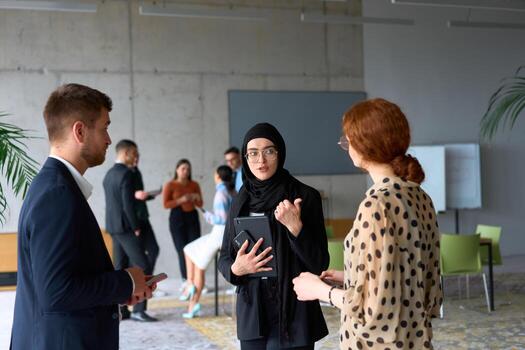 A diverse group of business professionals engages in a conversation during a work break in the office, with their colleagues visible in the background, fostering collaboration and connection photo