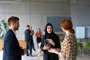 A diverse group of business professionals engages in a conversation during a work break in the office, with their colleagues visible in the background, fostering collaboration and connection photo
