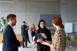 A diverse group of business professionals engages in a conversation during a work break in the office, with their colleagues visible in the background, fostering collaboration and connection photo