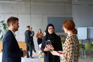 A diverse group of business professionals engages in a conversation during a work break in the office, with their colleagues visible in the background, fostering collaboration and connection photo
