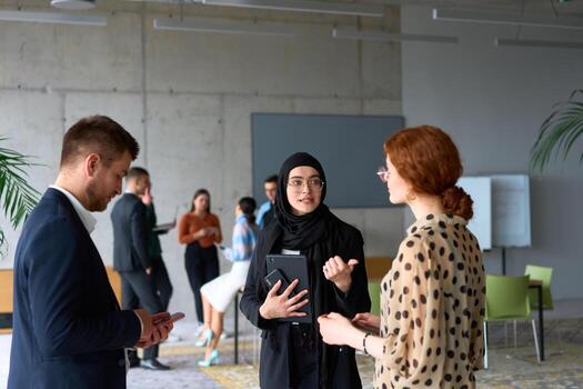 A diverse group of business professionals engages in a conversation during a work break in the office, with their colleagues visible in the background, fostering collaboration and connection photo