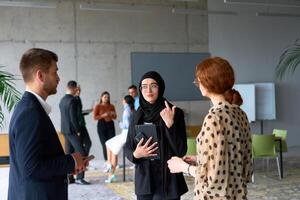 A diverse group of business professionals engages in a conversation during a work break in the office, with their colleagues visible in the background, fostering collaboration and connection photo