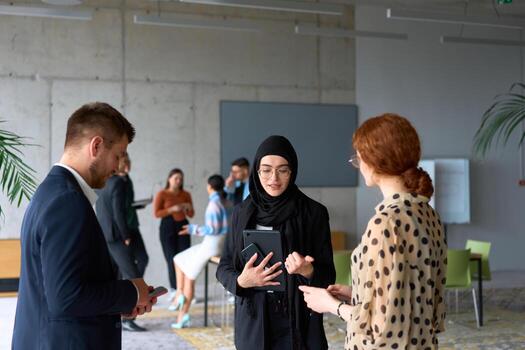 A diverse group of business professionals engages in a conversation during a work break in the office, with their colleagues visible in the background, fostering collaboration and connection photo