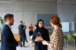 A diverse group of business professionals engages in a conversation during a work break in the office, with their colleagues visible in the background, fostering collaboration and connection photo