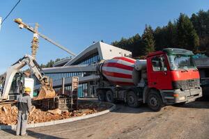 The exterior of a business building under construction is surrounded by trucks, cranes, and various construction materials, creating a bustling scene of an expansive industrial worksite in progress photo