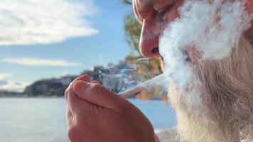 Close-up of a man with a gray beard lighting a cigarette against the background of the sea. bad habits. Thoughtful man smoking a cigarette with a filter. High-quality 4k footage video