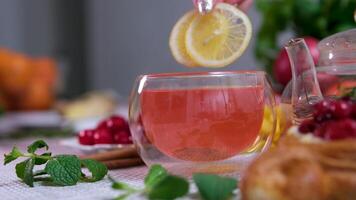 Two pieces of lemon in red hibiscus Pouring tea with orange, lemon and lime out of transparent tea pot into glass with nint.Brewing tea with honey and pancakes in background. Slow motion tea. video