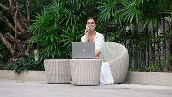 Beautiful woman sitting on a sofa, working remotely on a laptop while smiling and enjoying a relaxed, comfortable atmosphere. Freelancer smiling and working from green garden on laptop. video
