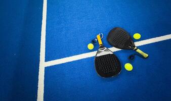 A padel or paddle tennis racket and a bright yellow tennis ball lie on a blue court. The net and a blurred figure of a player are visible in the background. photo