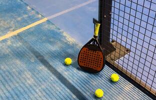 A padel or paddle tennis racket and a bright yellow tennis ball lie on a blue court. The net and a blurred figure of a player are visible in the background. photo