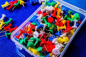 Colorful push pins in plastic box on blue background, office supplies close up photo
