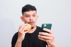 an asian man is enjoying his hamburger while holding his smartphone photo