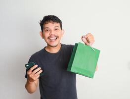 portrait of happy smiling handsome Asian man carrying shopping bags and showing smartphone, online shopping concept photo