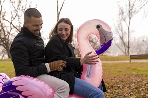 portrait of two young lovers having fun riding an inflatable pink flamingo in a public park in winter photo