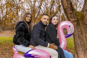 small group of young friends having fun riding an inflatable pink flamingo over a wooden table in a public park in winter photo