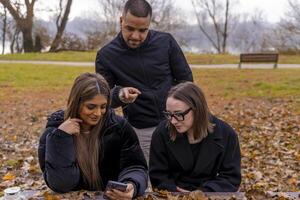 small group of young friends having fun using smart phone sitting on a wooden table in a public park in winter photo