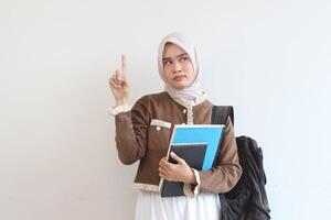 A Young Muslim Woman Student Holding Notebooks and Backpack, Pointing Upward an Idea photo