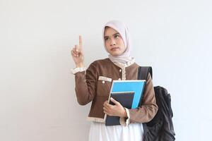 A Thoughtful Muslim Student Girl Holding Notebooks and Backpack Against White Wall photo