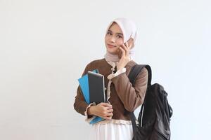 A Smiling Muslim Female Student on a Phone Call, Holding Notebooks and a Backpack Against a White Background photo