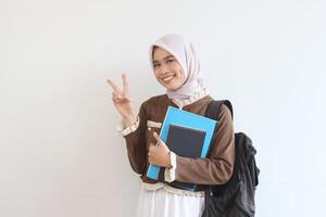 Smiling Muslim Student Girl Holding Books and Backpack Showing Peace Sign Against White Background photo