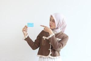 A Young Woman in a Brown Jacket and Pink Hijab Shows a Blue Credit Card Against a White Background photo