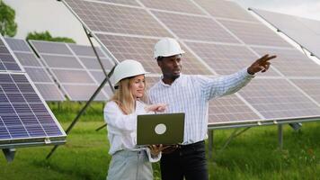 Diverse female and male engineers in hardhats using laptop and discussing project while standing near photovoltaic batteries in countryside video