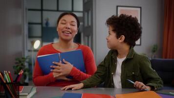 African American family at home studying education together personal teacher woman girl female mother holding book textbook talking to little child kid boy son touch mom nose with pen learning help video