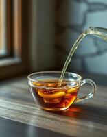 Warm tea being poured into a clear glass cup on a wooden table in a cozy setting at afternoon photo