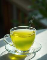 Hot green tea steaming in a clear glass cup on a sunlit table with soft shadows photo