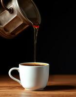 Rich coffee brewed and poured from a metal pot into a white cup on a wooden table against a dark background photo