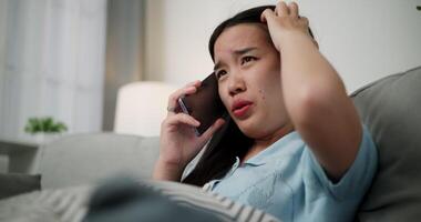 Handheld low angle view shot, An upset young woman is talking on the phone to someone with an angry and irritated look on her face while sitting on the sofa in living room at home. video